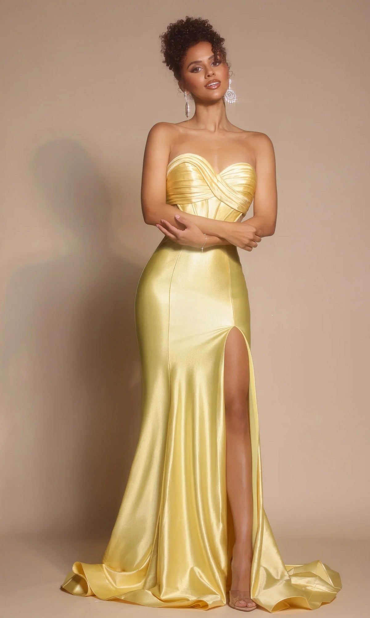 A woman models the Portia and Scarlett XO066 long formal dress in yellow, featuring a strapless satin design with a pleated crossover bodice, sweetheart neckline, and thigh-high slit, set against a beige background.